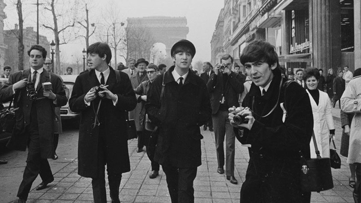 Paul, John y George caminan por las calles de París, con el Arco del Triunfo de fondo, rodeados por fotógrafos y franceses sorprendidos 