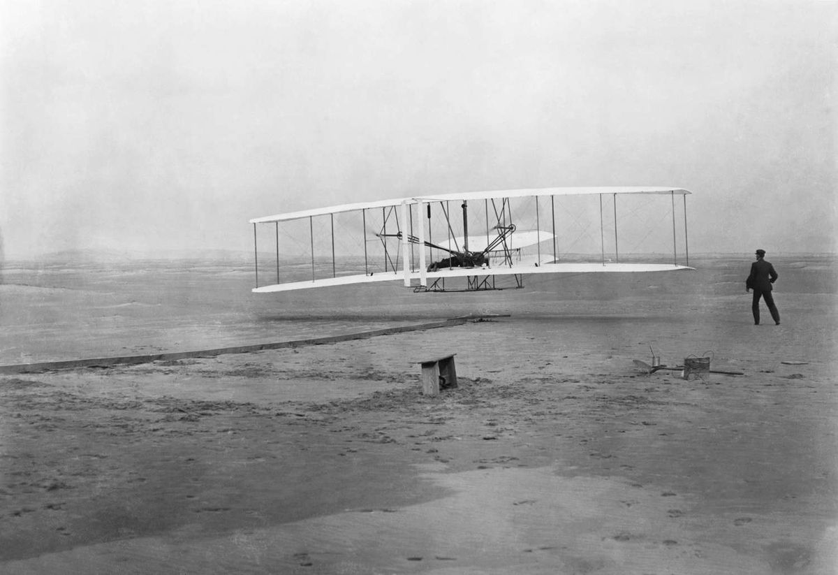 El histórico primer vuelo del 17 de diciembre de 1903, con Orville Wright acostado boca abajo sobre el ala inferior, y con las caderas en el soporte que operaba el mecanismo de deformación del ala, y su hermano Wilbur corriendo al lado de la máquina. Detrás de la misma, se observan el riel de arranque, el apoyo del ala, una caja de bobinas y otros elementos necesarios para la preparación del vuelo. El histórico primer vuelo del 17 de diciembre de 1903, con Orville Wright acostado boca abajo sobre el ala inferior, y con las caderas en el soporte que operaba el mecanismo de deformación del ala, y su hermano Wilbur corriendo al lado de la máquina. Detrás de la misma, se observan el riel de arranque, el apoyo del ala, una caja de bobinas y otros elementos necesarios para la preparación del vuelo.